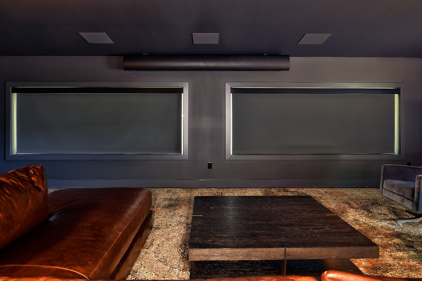 A wide image of a home theater room with motorized shades over the windows and a comfy leather couch.